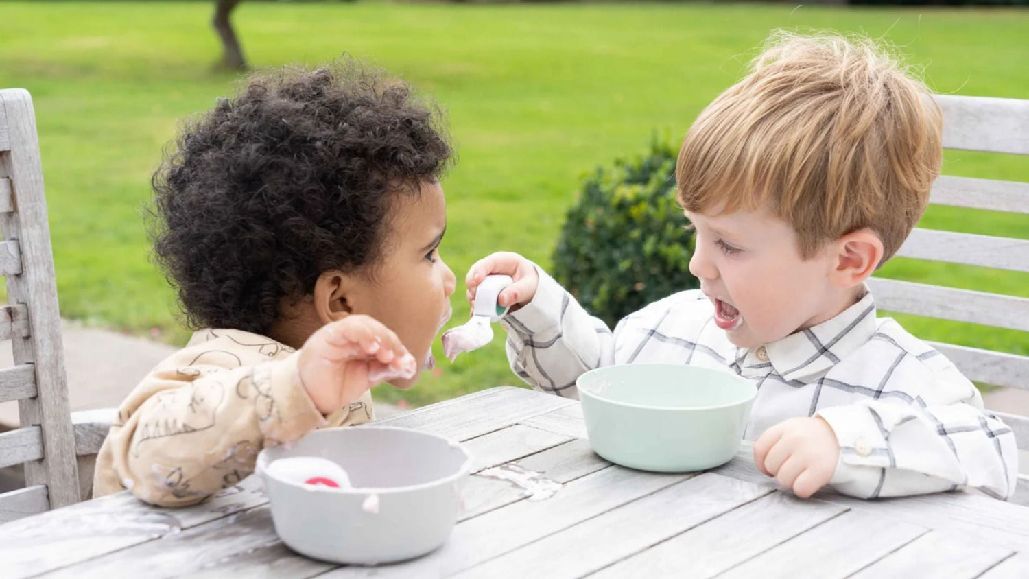 Two toddler boys sit at a garden table sharing yoghurt
