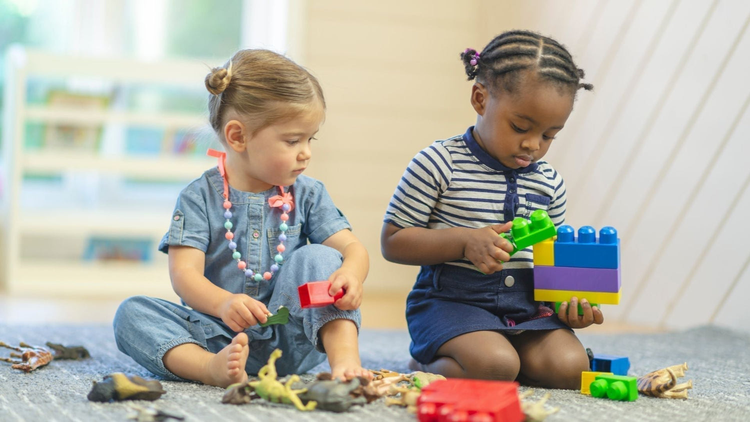 two toddlers sit on carpet playing side by side with blocks