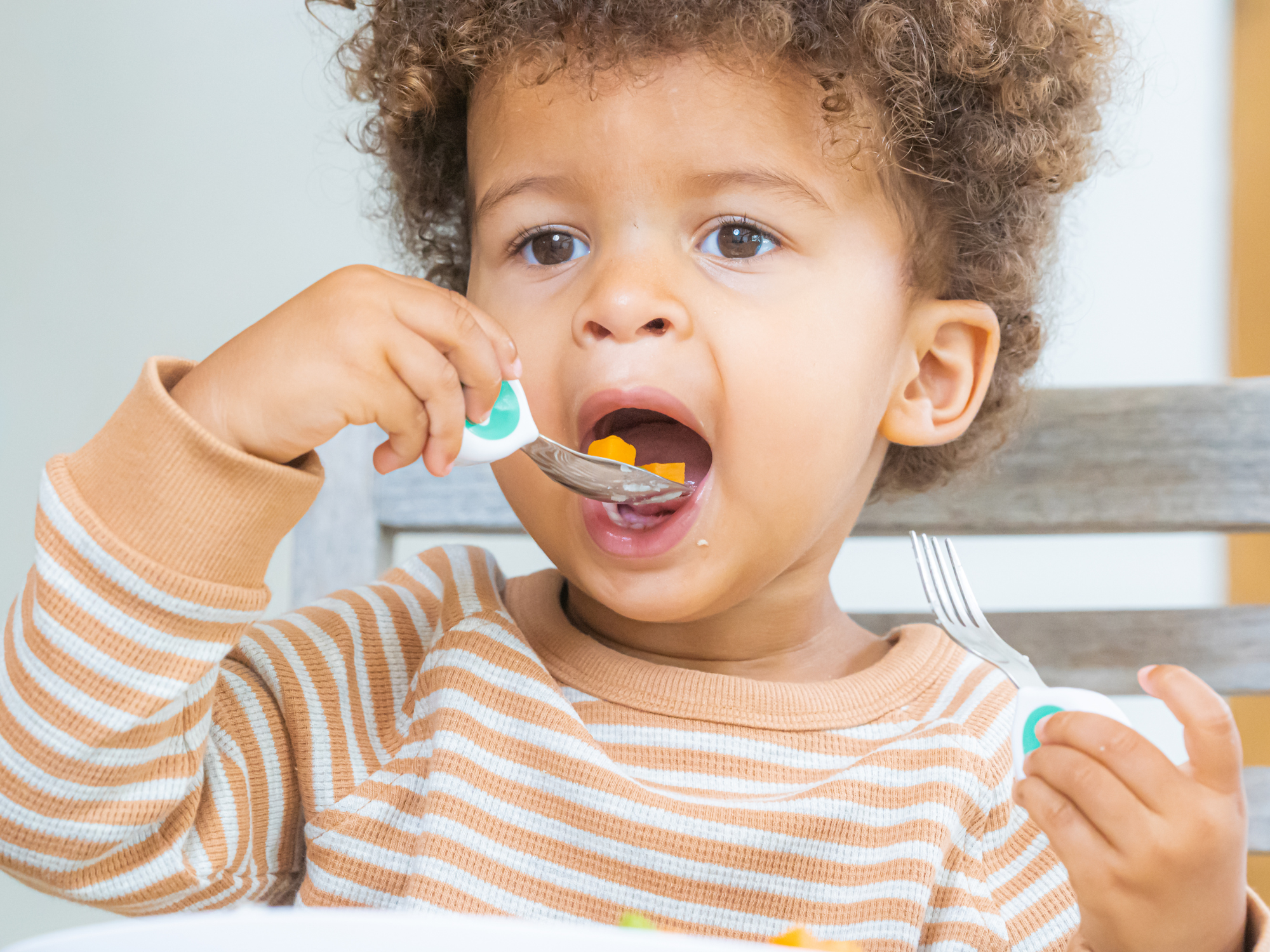 introducing doddl toddler cutlery. a toddler boy in a stripy t-shirt uses a doddl spoon to spoon food into his mouth