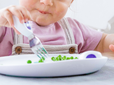 A toddler spearing peas on a white plate with a doddl fork