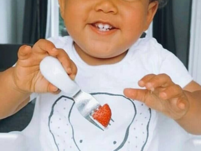 A smiling toddler holding a doddl fork with a strawberry on the end