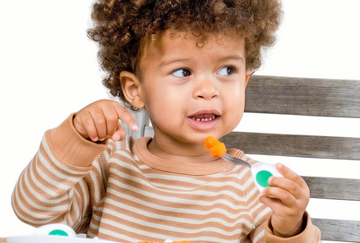 Child sitting at a table with a plate of food, using a spoon and fork.