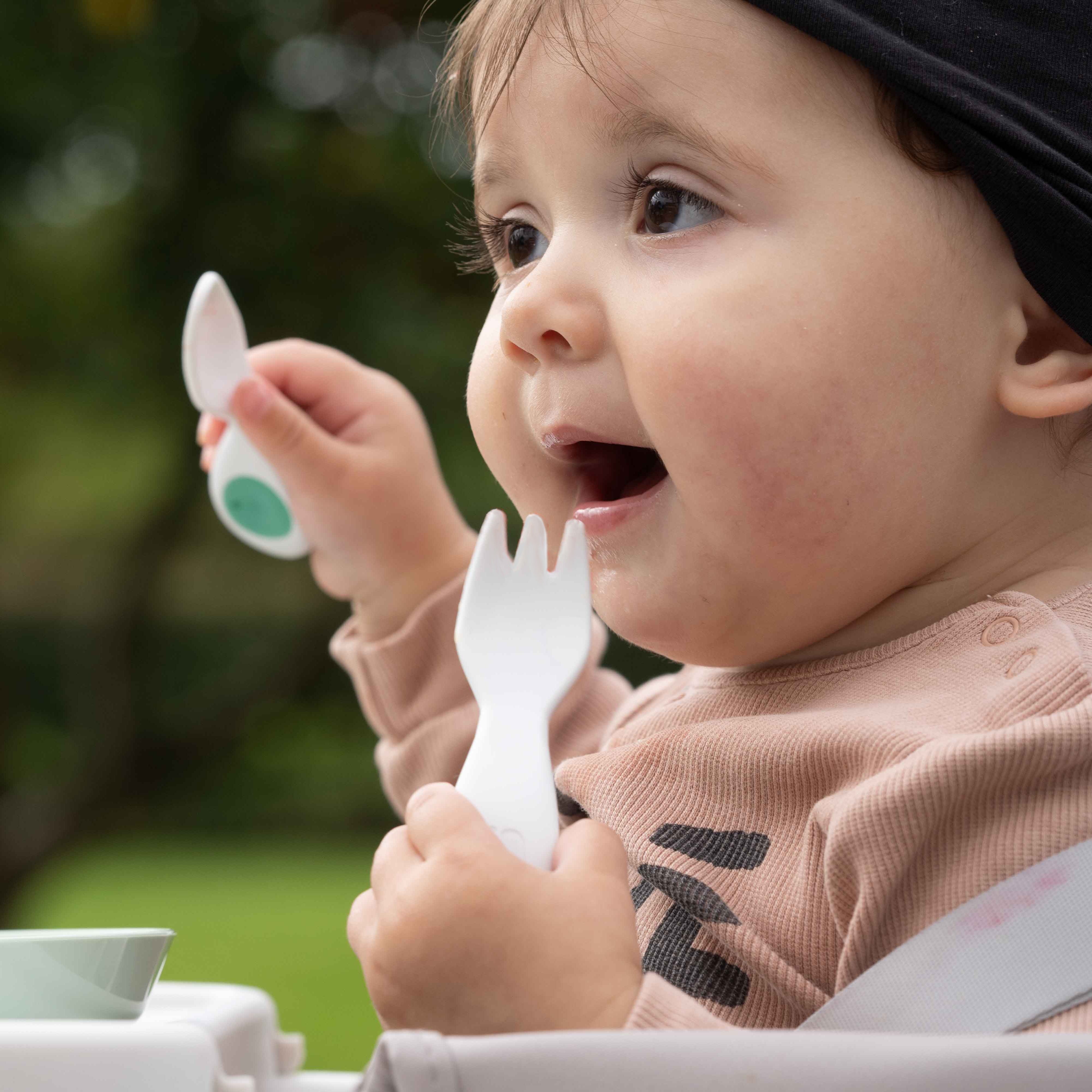 A close-up lifestyle shot of a baby sitting in a high chair outdoors, holding a white plastic doddl fork with a green circular accent. The baby is wearing a light pink top and a black head covering, with a blurred green garden background.