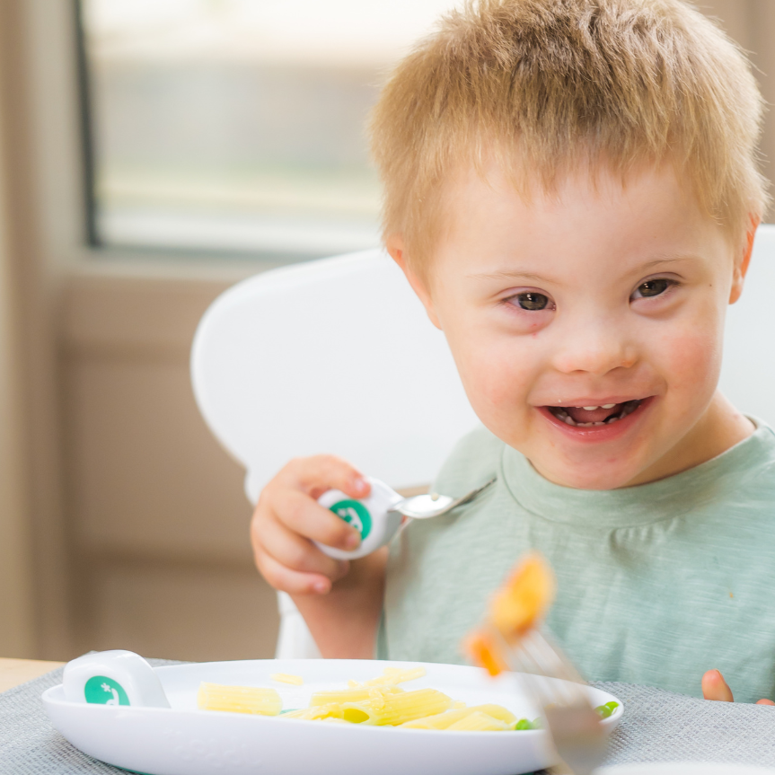 A young child with down syndrome smiling while sitting in a white high chair, holding a doddl spoon with a green handle, in a brightly lit indoor setting.