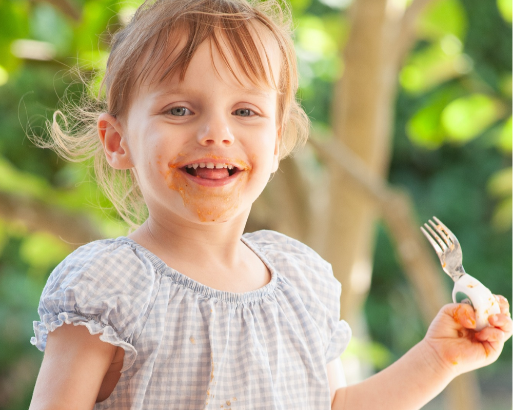 An autistic young girl with food smeared on her face smiles while holding a doddl fork outdoors, wearing a light blue and white gingham dress against a blurred green garden background.