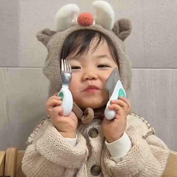 Child wearing a bear hat holding a doddl fork and spoon against a gray tiled wall.