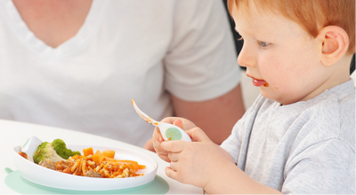 A toddler with red hair wearing a light gray shirt sits at a table, holding a doddl baby spoon and looking at a plate of food with broccoli and carrots, while an adult in a white shirt is seated behind them.