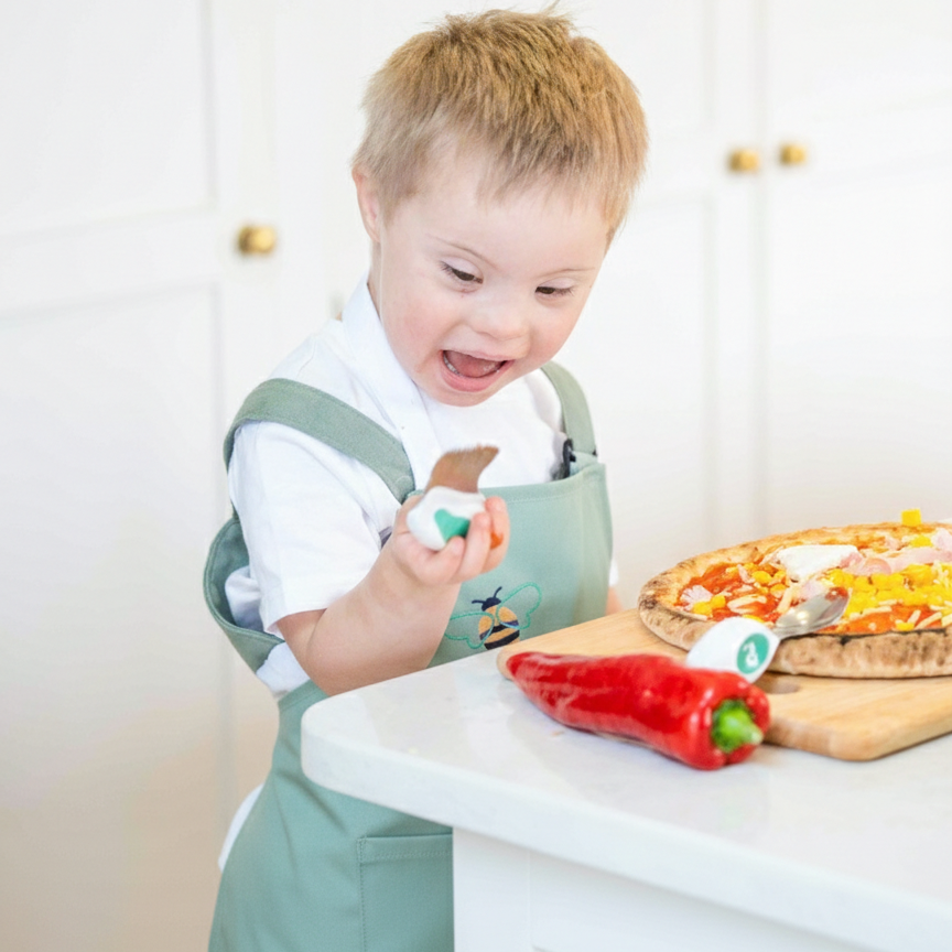 Child in a green apron chopping with doddl knife - pizza and pepper on a kitchen counter.