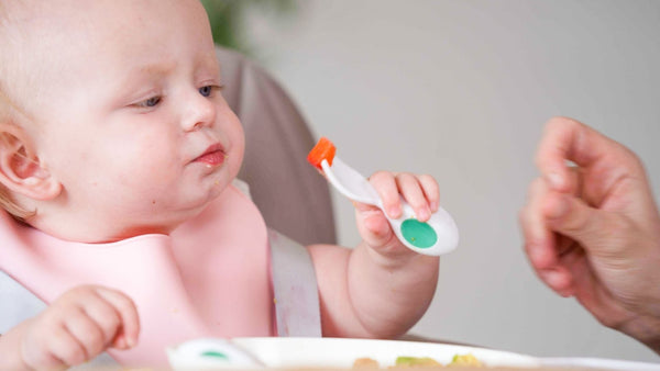 Baby sits in a highchair using a doddl baby spoon to feed themselves, the image shows the grip of the hand on the spoon