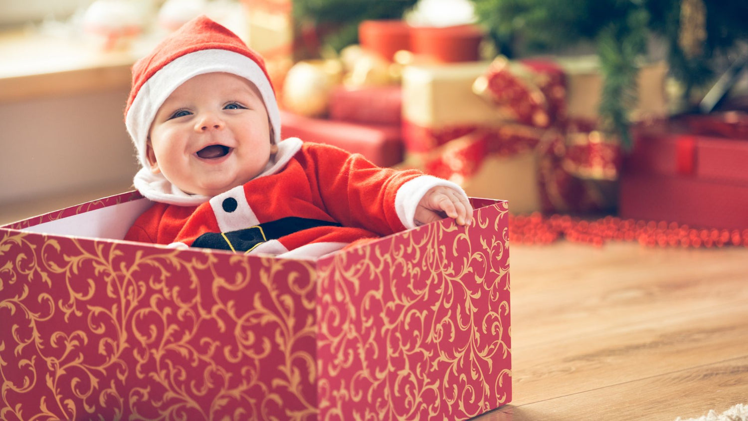 Happy smiling toddler in Santa outfit in a gift box by a Christmas tree