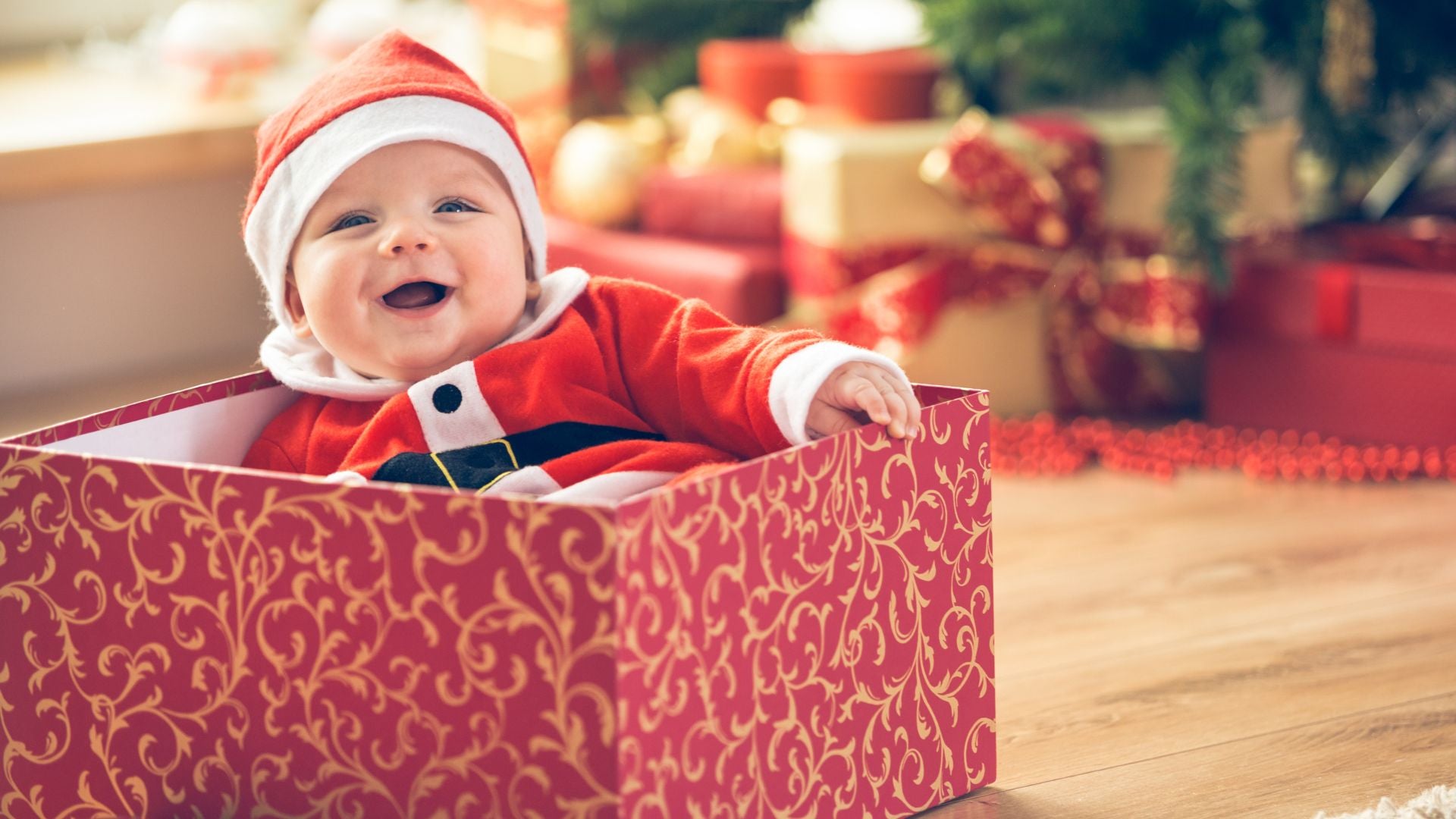 Happy smiling toddler in Santa outfit in a gift box by a Christmas tree