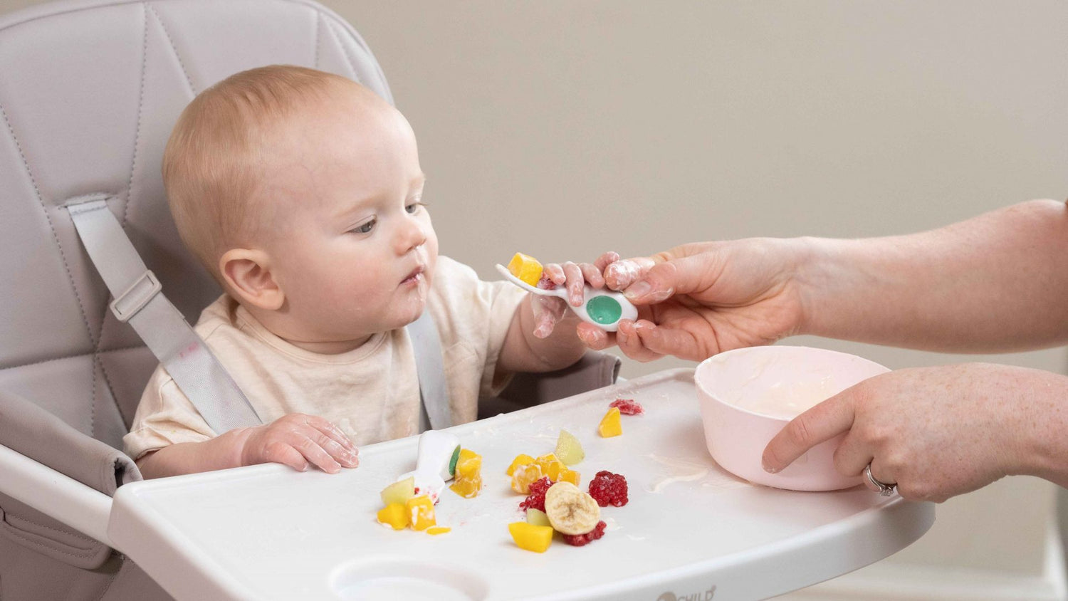 baby boy sits in highchair, being handed a doddl spoon with fruit in