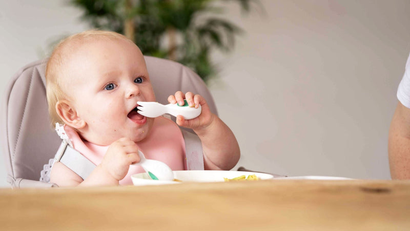 Happy baby sitting at a table in a highchair, having first tastes of food with doddl baby cutlery