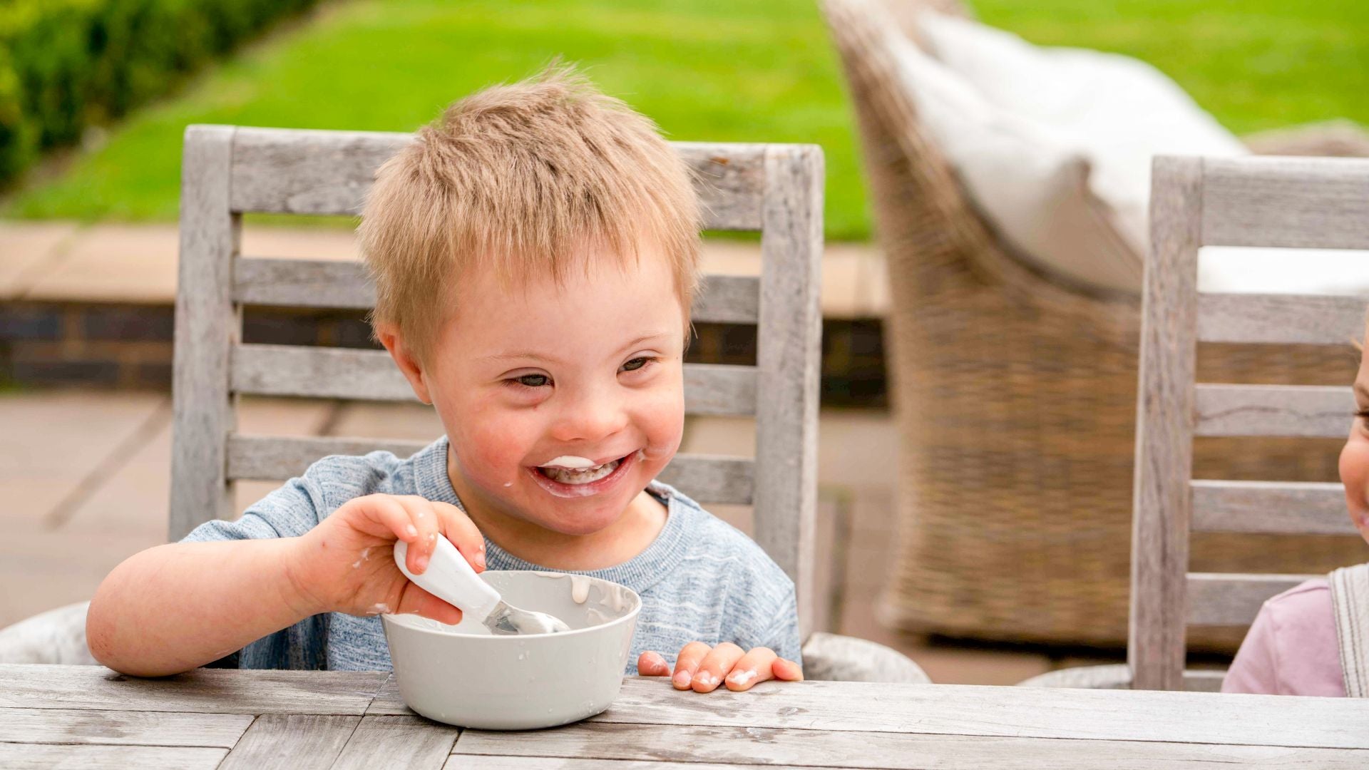 Toddler boy sits at a garden table, he is smiling and eating yoghurt from a bowl with his doddl toddler spoon
