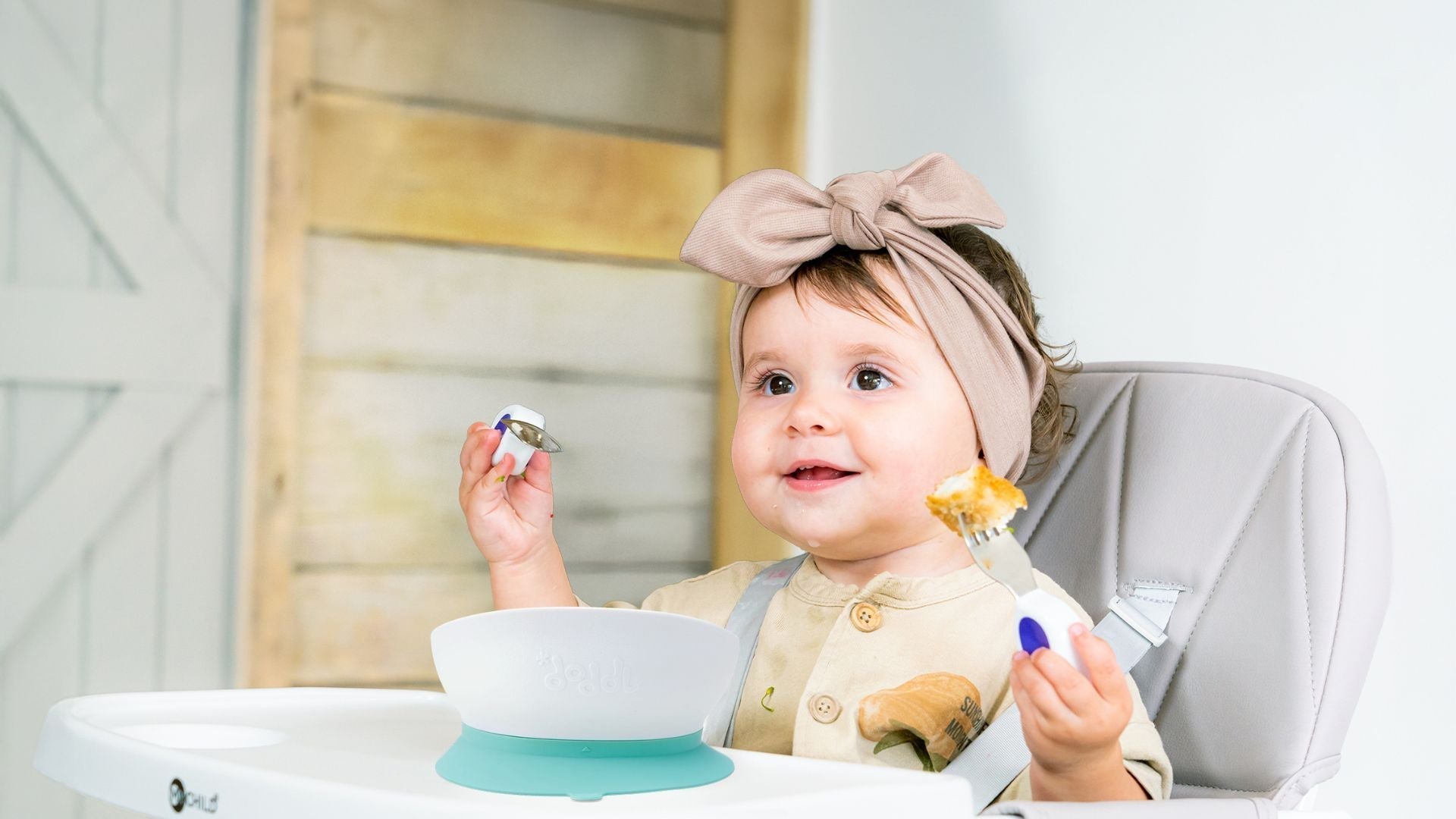 Happy toddler girl eating at a table with doddl fork, spoon and bowl
