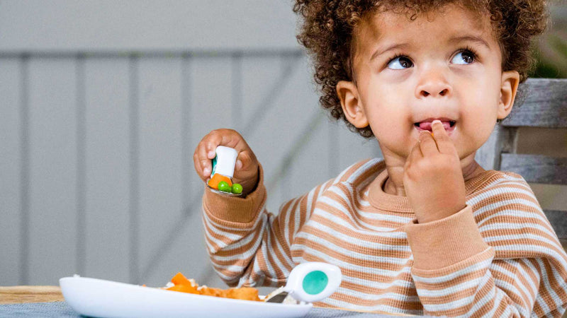 toddler boy sitting at a meal table eating with a doddl fork and spoon on a doddl plate