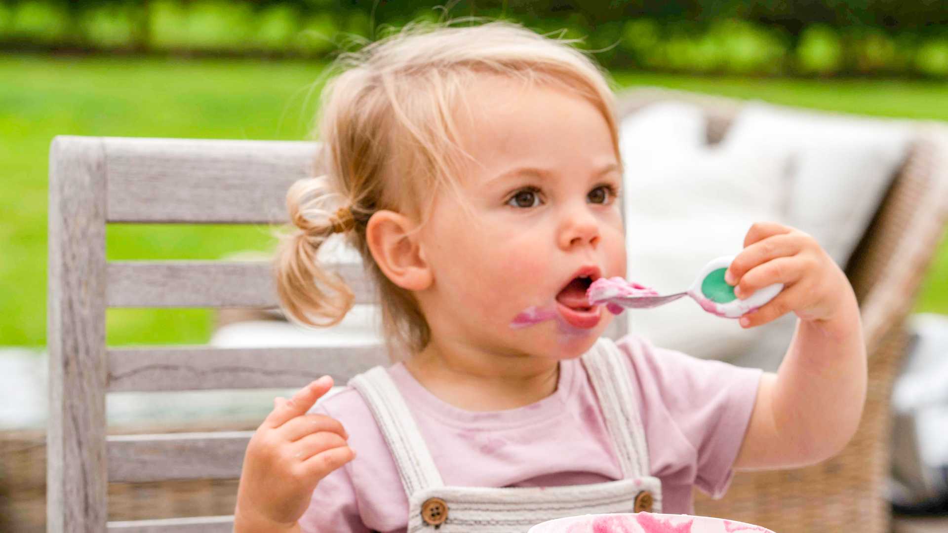 Toddler girl eating pink yoghurt with a doddl toddler spoon in a garden