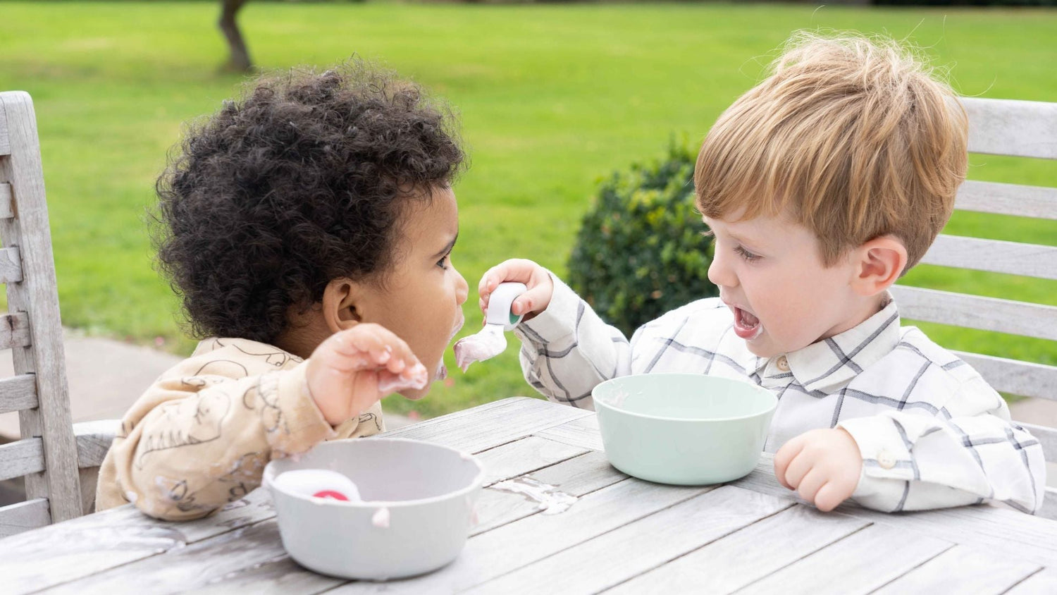 Two toddler boys sit at a garden. They both have bowls of yoghurt, one uses his toddler spoon to feed the other