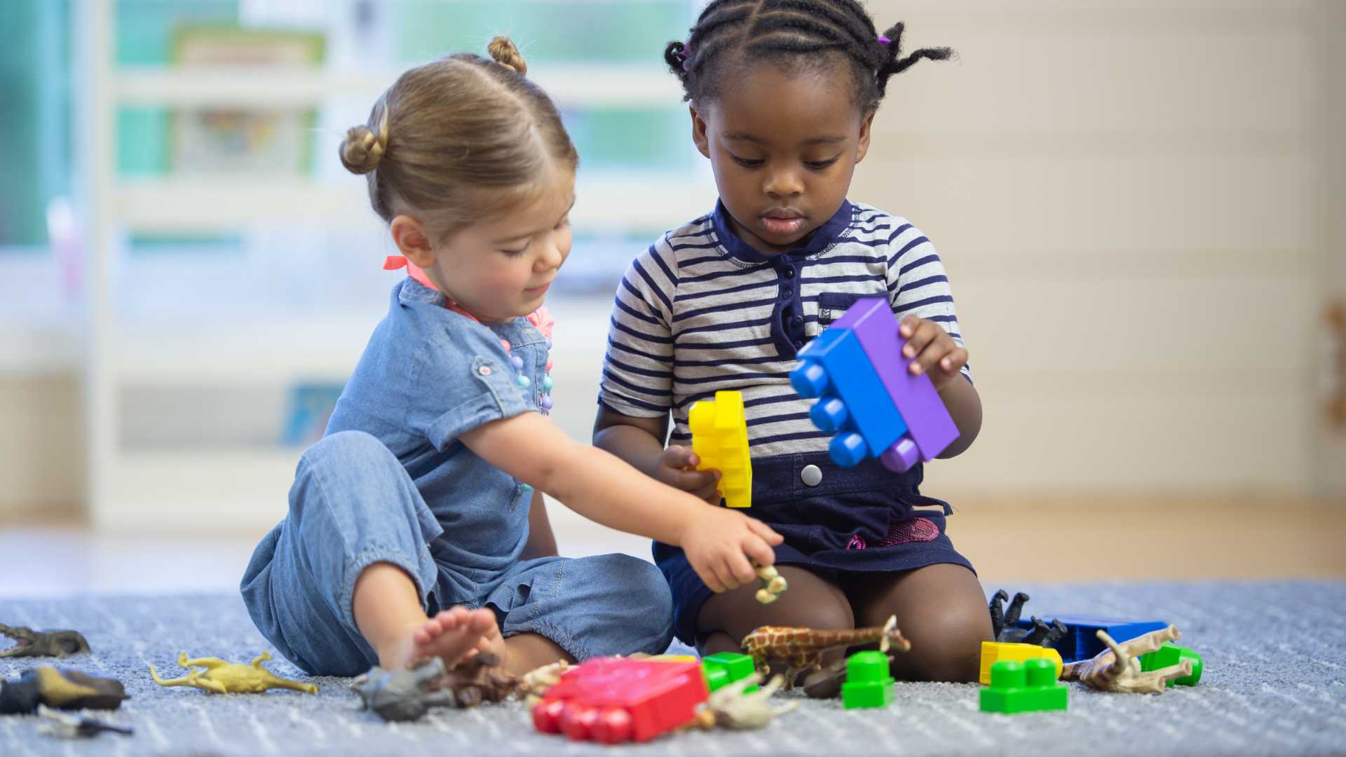 two toddlers sit playing together on the floor using blocks to build a tower