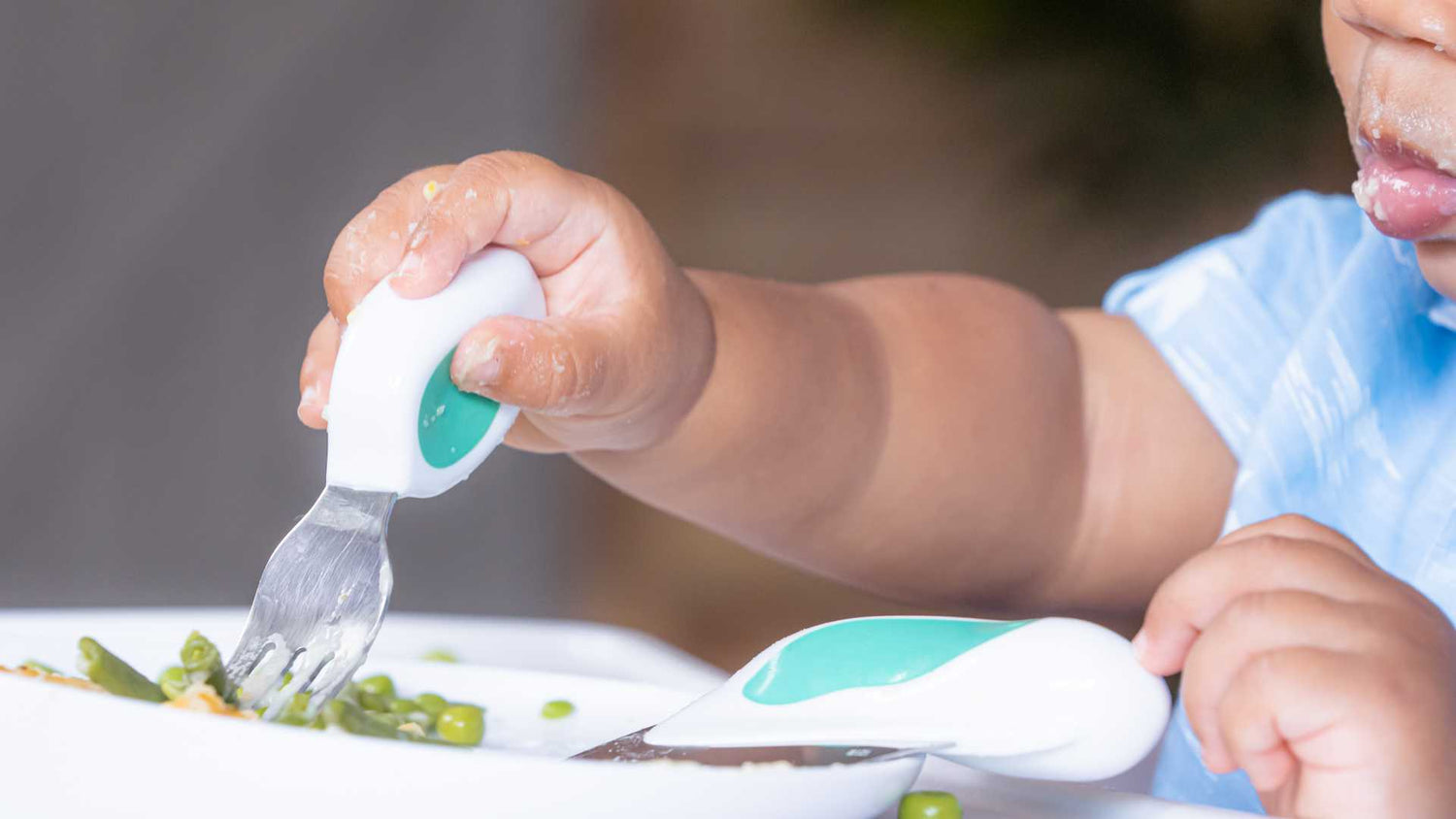 close up of toddler arm and hand holding a doddl fork to eat peas from a plate