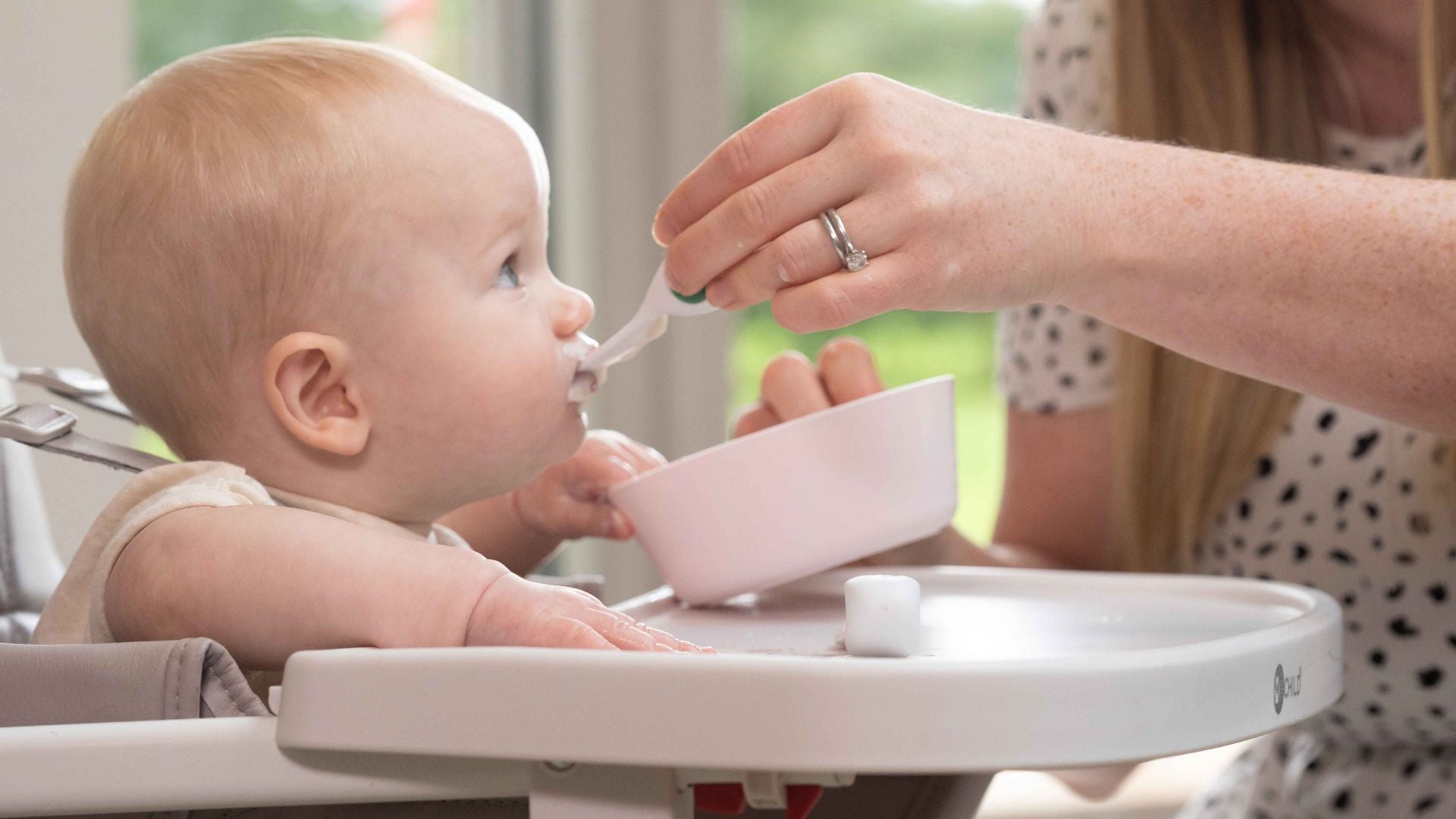 baby sits in a highchair being fed by his mum, with a doddl baby spoon