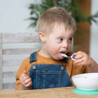 A young child with Down syndrome sitting at a wooden table, wearing blue denim overalls and an orange t-shirt, eating from a white bowl with a purple-handled doddl spoon.