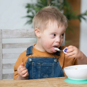 A young child with Down syndrome sitting at a wooden table, wearing blue denim overalls and an orange t-shirt, eating from a white bowl with a purple-handled doddl spoon.