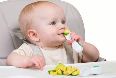 Baby sitting in a high chair eating with a doddl spoon and showing avocados.