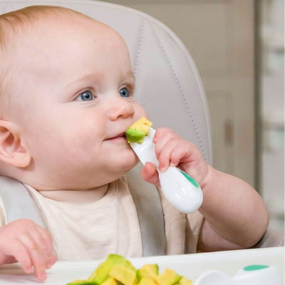 Baby in a high chair eating with a doddl 
spoon and showing avocados on a plate.