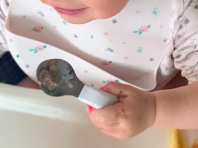 A toddler in a floral bib holding a doddl spoon of food in a whole-hand grip