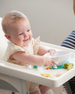 Baby in a high chair smiling while self-feeding with doddl baby cutlery