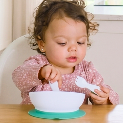 Child sitting at a table with doddl bowl and utensils, wearing a pink floral shirt.