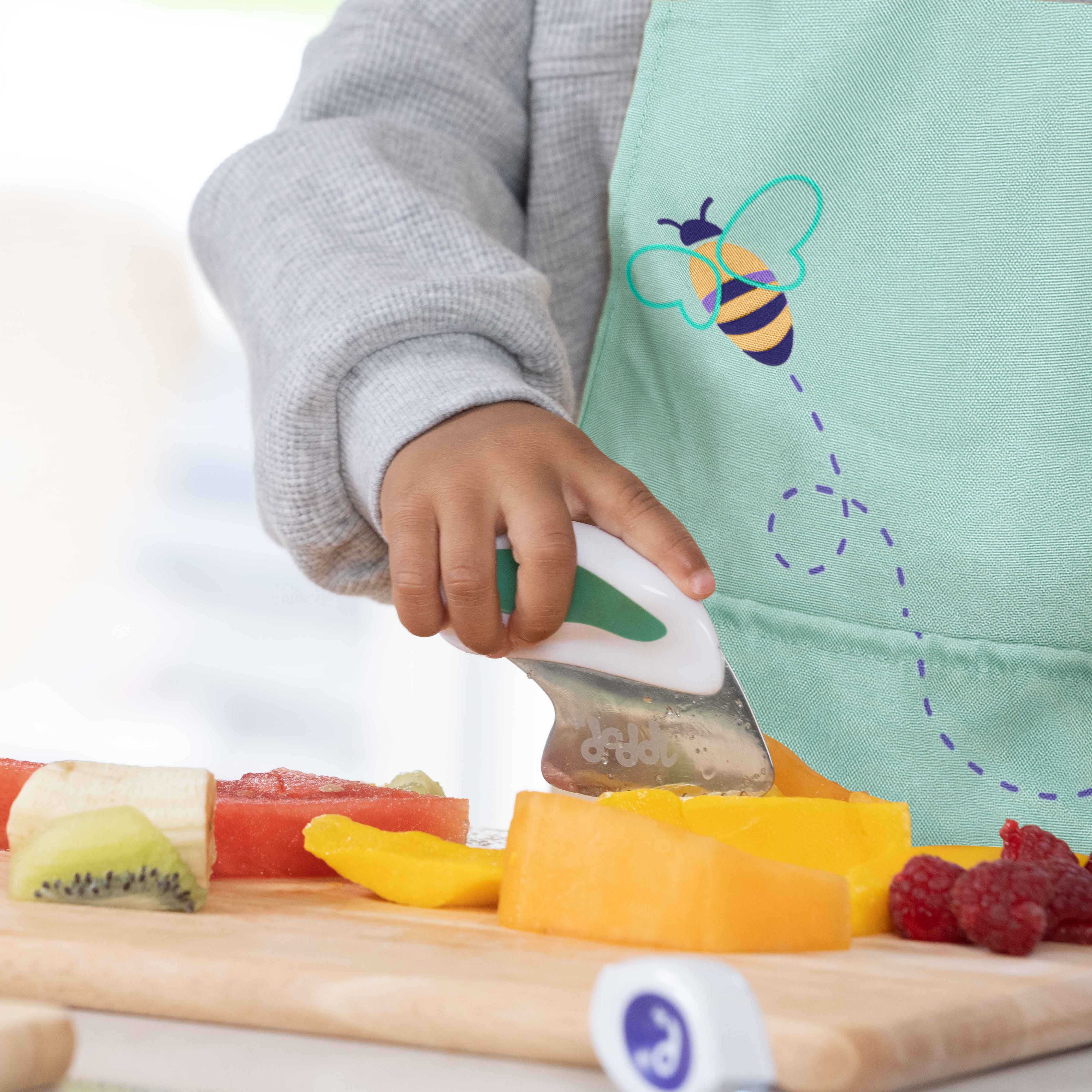Child cutting fruits on a wooden board with a knife, wearing a green apron with a bee design.