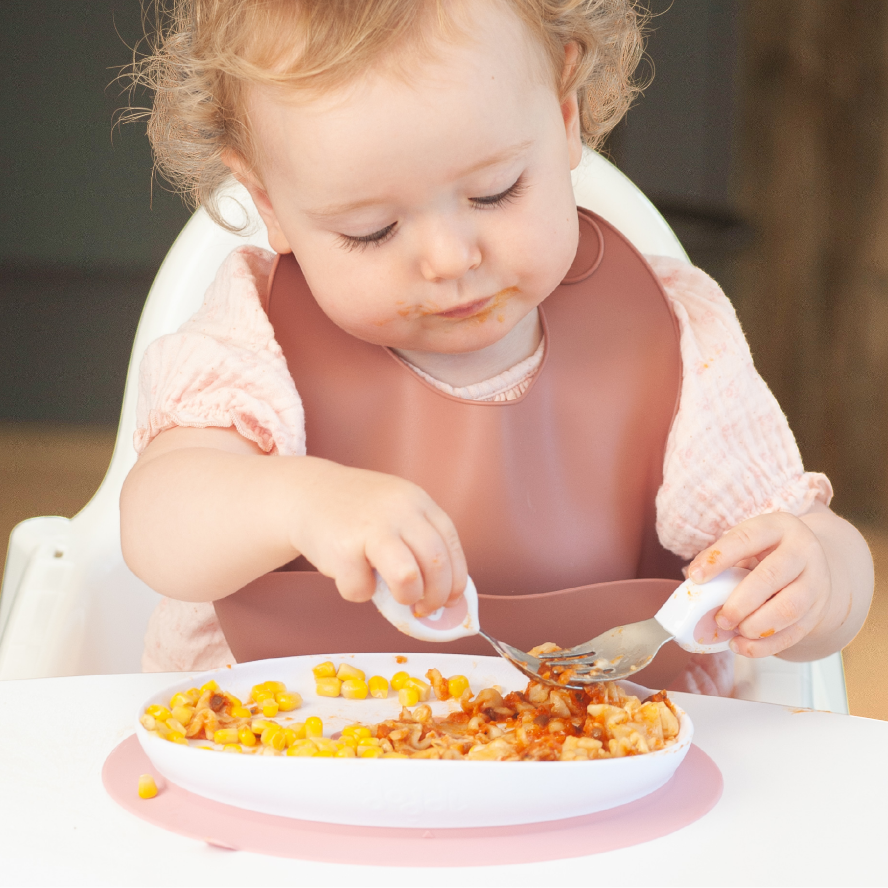 A toddler with curly blonde hair sits in a white high chair, wearing a dusty rose pink bib and eating from a white doddl plate with a doddl spoon and doddl fork.