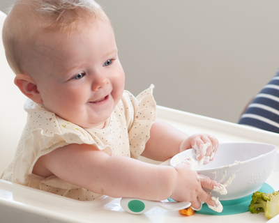 A baby sitting in a white high chair, wearing a cream polka-dot bib and interacting with a doddl bowl on a teal base. The baby's hands and face are covered in food, with broccoli and other pieces visible on the tray.