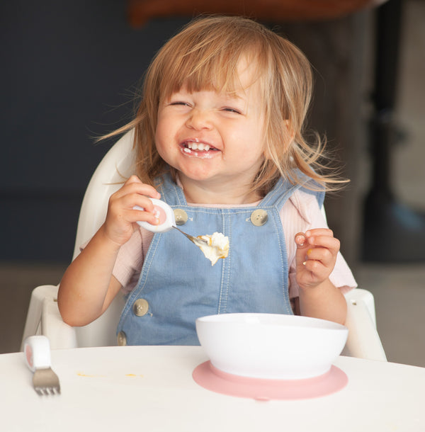 A young toddler with blonde hair sits in a white high chair, smiling while holding a doddl spoon with food. The child is wearing a light blue denim overall dress over a pink shirt.