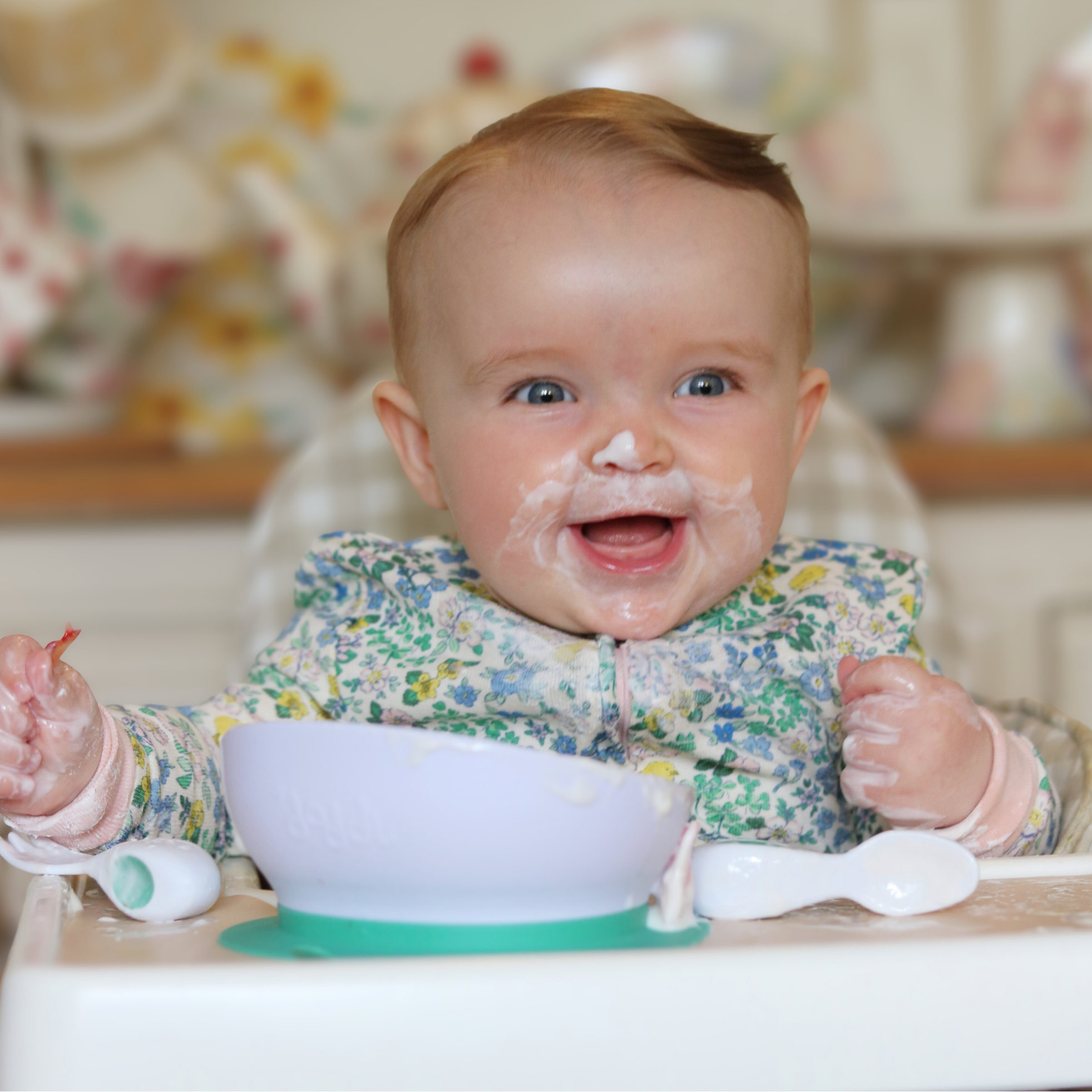 Baby in a high chair with a doddl bowl and spoon, smiling with food on face
