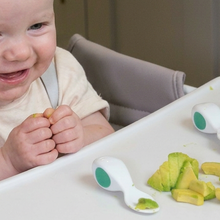 A baby smiling in a high chair, with two white baby spoons featuring green circular accents placed on the tray next to cut avocado pieces.