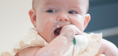 A close-up lifestyle shot of a baby sitting in a high chair, covered in white food residue, with a white doddl bowl in the foreground and a soft-focus background.