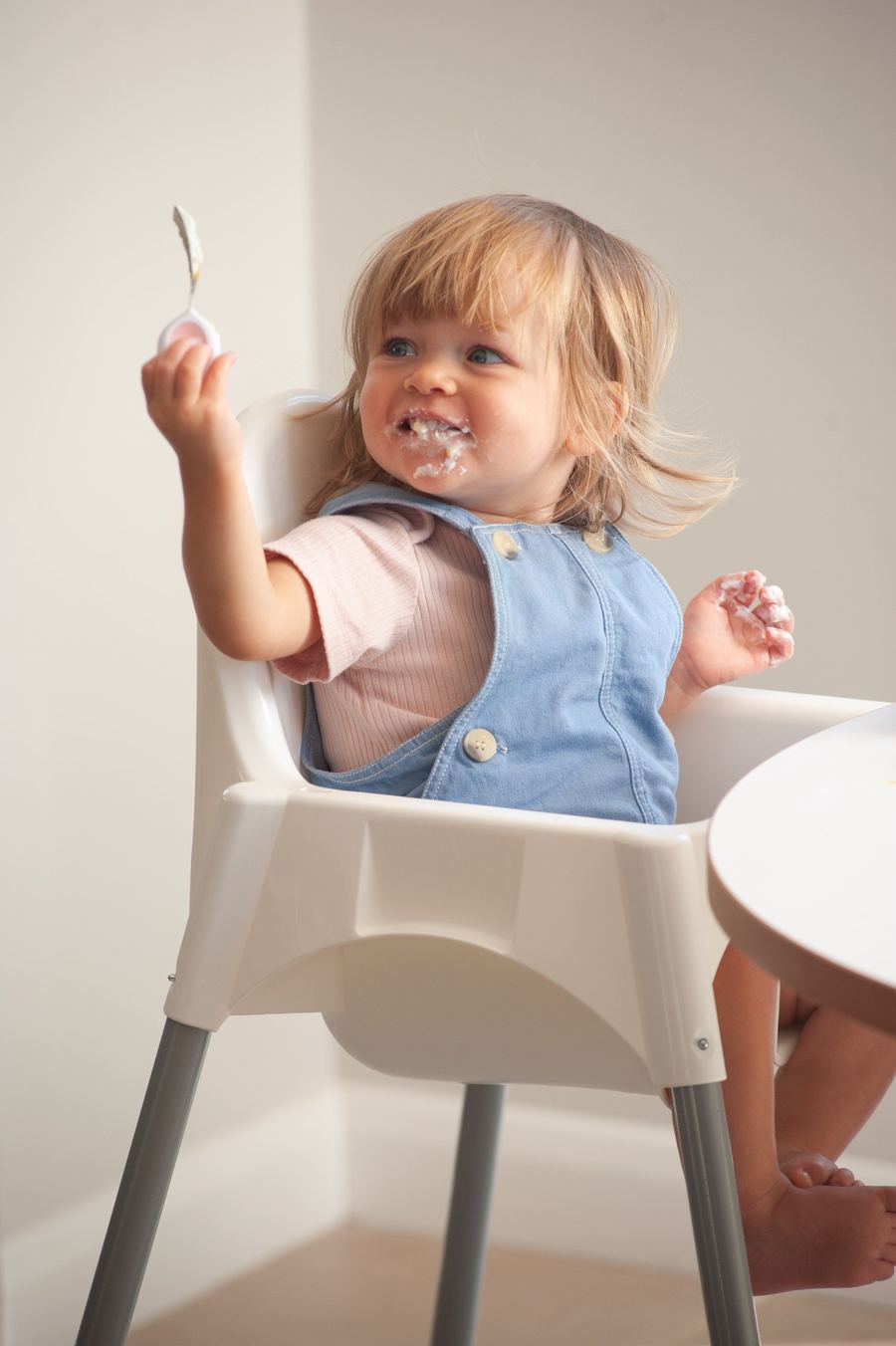 A smiling toddler in a high chair holding up a doddl spoon after eating